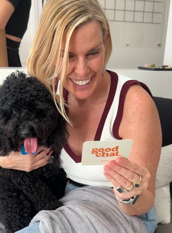 Smiling woman with a black poodle holding up a Good Chat card in bright indoor light