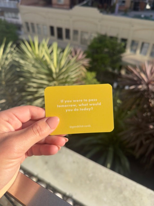 Yellow Good Chat Death deck conversation card held up in bright daylight over rooftop palms