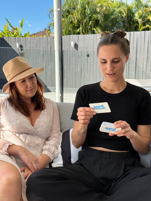 Three friends on an outdoor couch playing Good Chat conversation cards, one reading a question card aloud