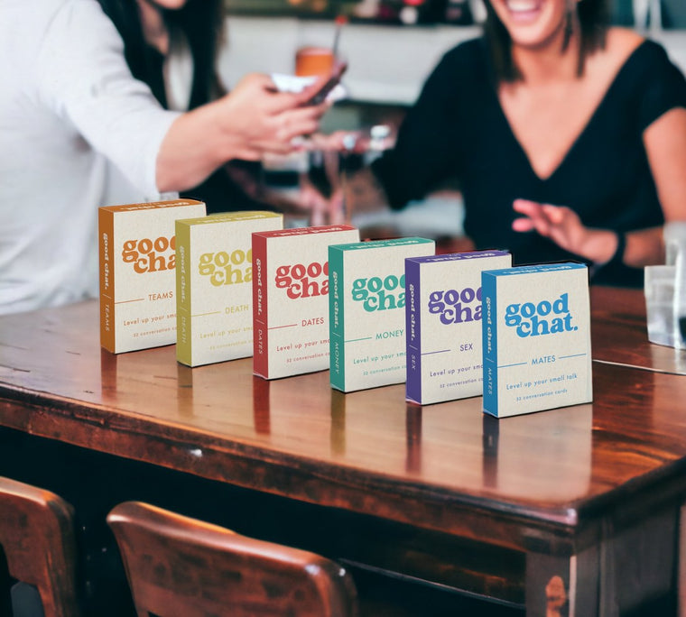 All six Good Chat decks lined up on a table with friends toasting behind