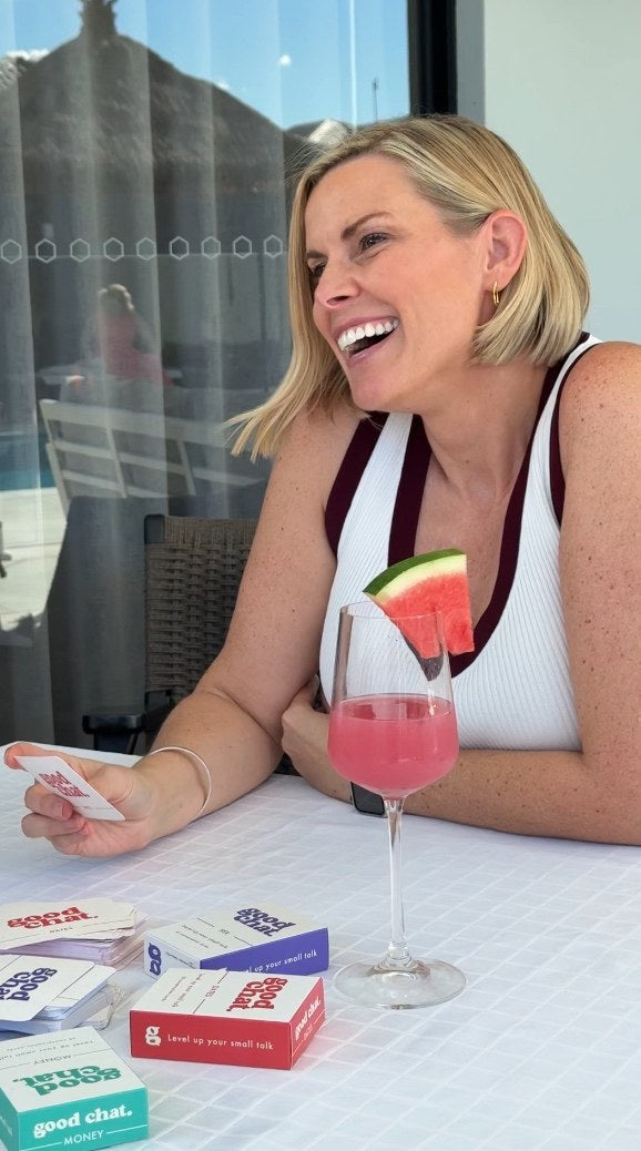 Laughing woman with a watermelon cocktail holding up a Good Chat card at a white table