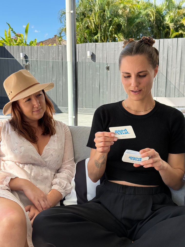 Three friends on an outdoor couch playing Good Chat conversation cards, one reading a question card aloud