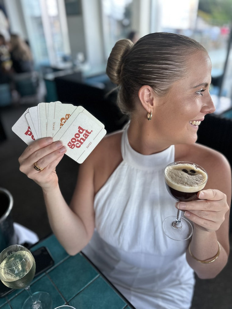 Woman fanning Good Chat cards in one hand with an espresso martini in the other at a bright bar