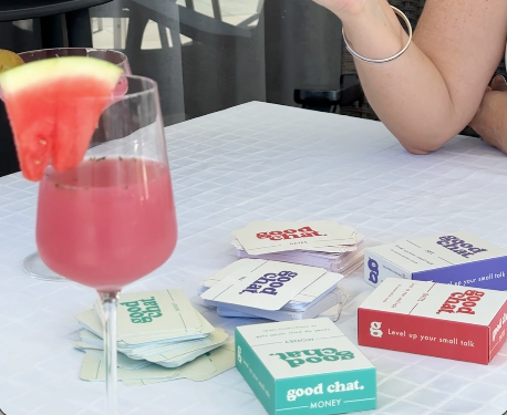Pink cocktail with watermelon slice and 'Good Chat' conversation cards on a table